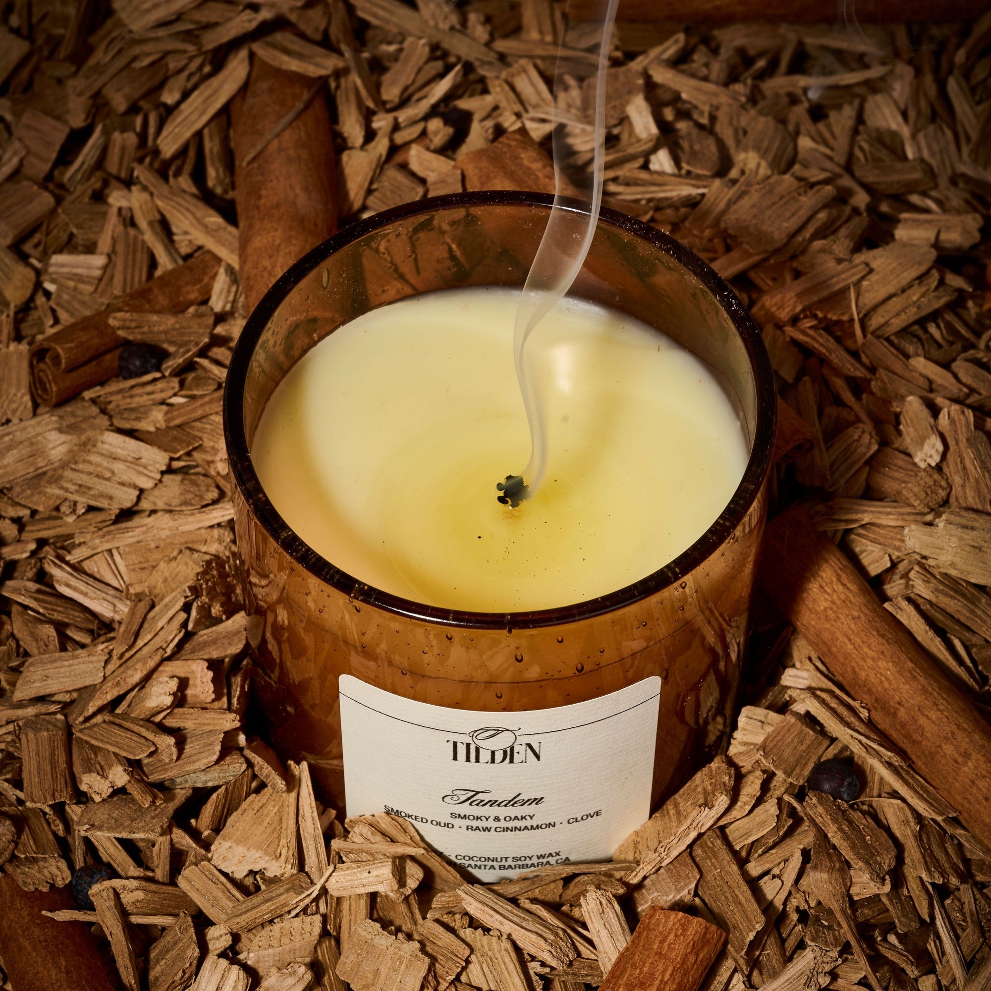 Candle in a wooden container on a bed of wood chips with cinnamon sticks.