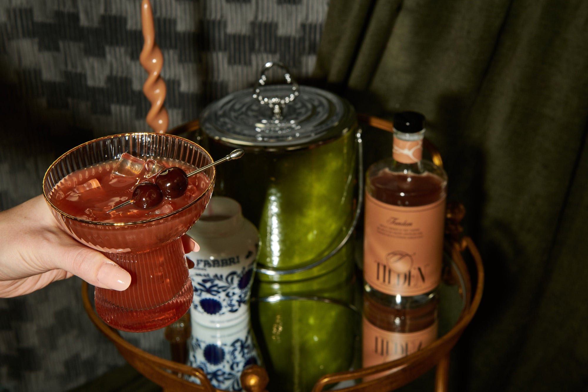 Person holding a non-alcoholic cocktail with a bar cart featuring bottles and glasses.